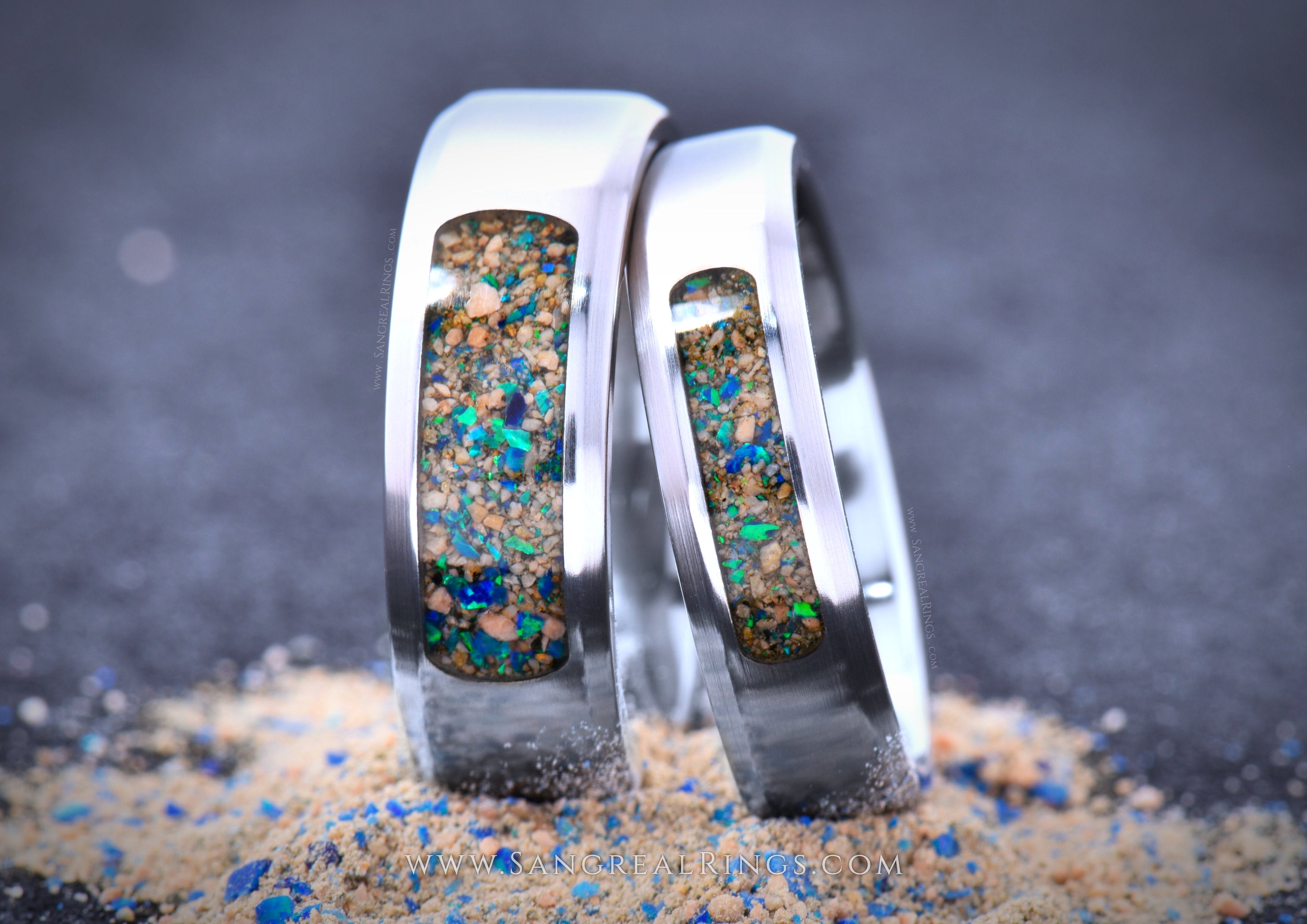 Two silver colored Titanium wedding rings with embedded colorful opal and stones on a sandy surface.