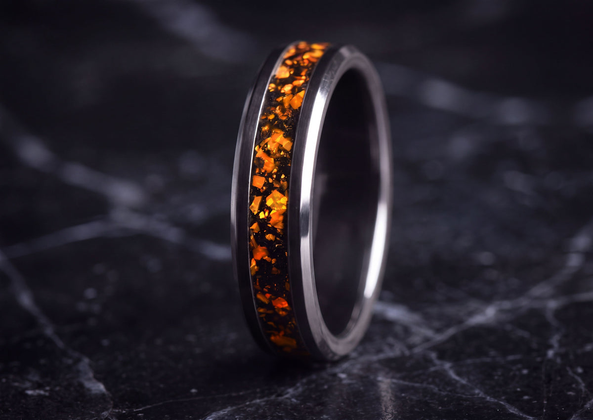 A polished dark metallic ring with a vibrant orange and black textured inlay, standing upright on a dark marble surface.