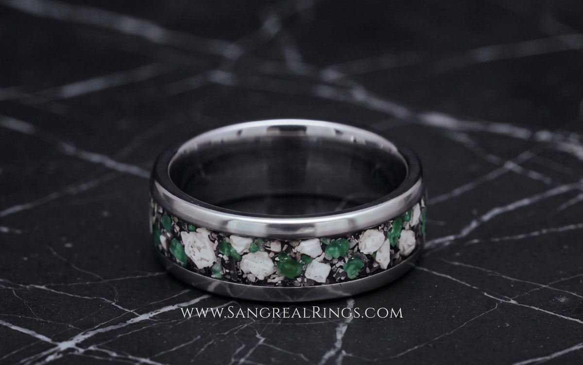 A silver-toned titanium band ring with a channel inlay of green emarald and white cremation ashes, displayed on a dark marble surface.