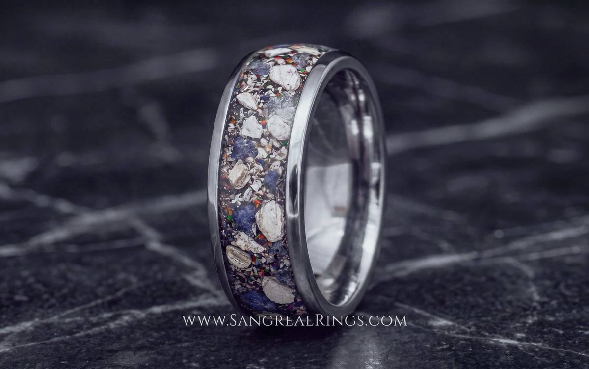 A polished silver-toned titanium ring with a textured inlay of small cremation ashes shards and blue sapphire shards, standing upright on a dark marble surface.