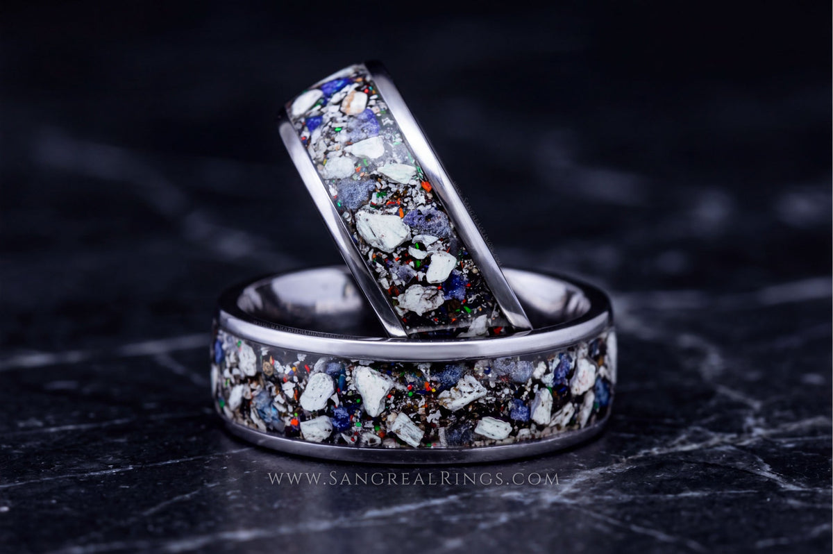 Two silver rings with a multicolor inlay of white, blue, and orange flecks, displayed on a dark textured surface.