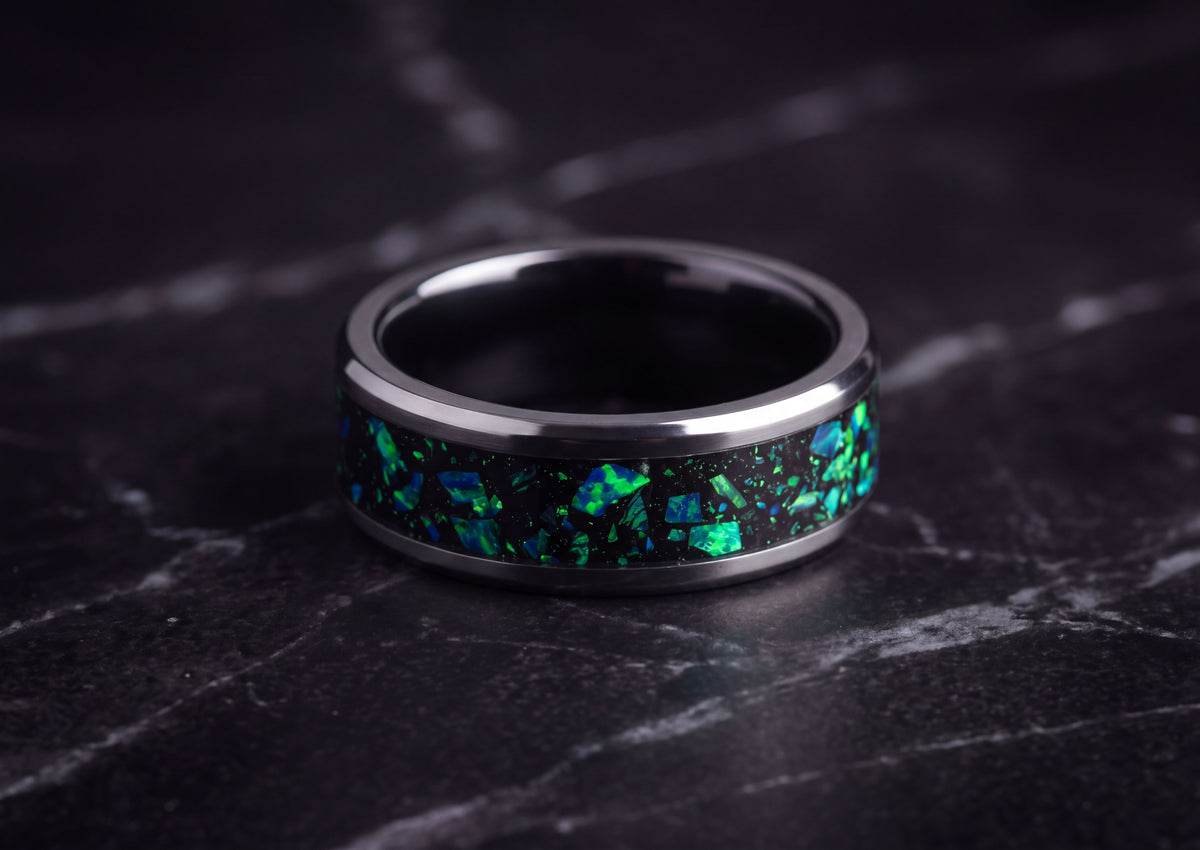 A polished dark ceramic ring band with a black inlay featuring vibrant green and blue iridescent flecks, resting on a dark marble surface.
