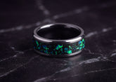 A polished dark ceramic ring band with a black inlay featuring vibrant green and blue iridescent flecks, resting on a dark marble surface.