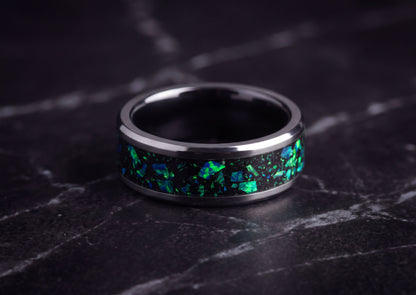 A polished dark ceramic ring band with a black inlay featuring vibrant green and blue iridescent flecks, resting on a dark marble surface.