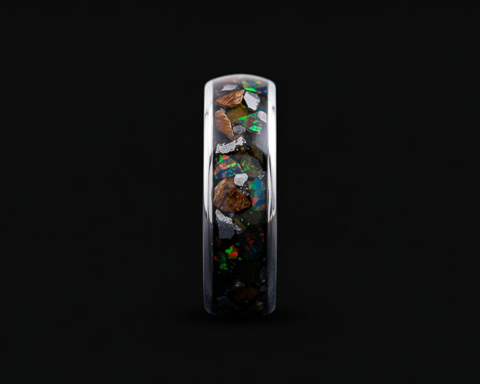 A polished silver band ring featuring an inlay of dark volcanic rock and vibrant, multicolored opal fragments, shown against a solid black background.