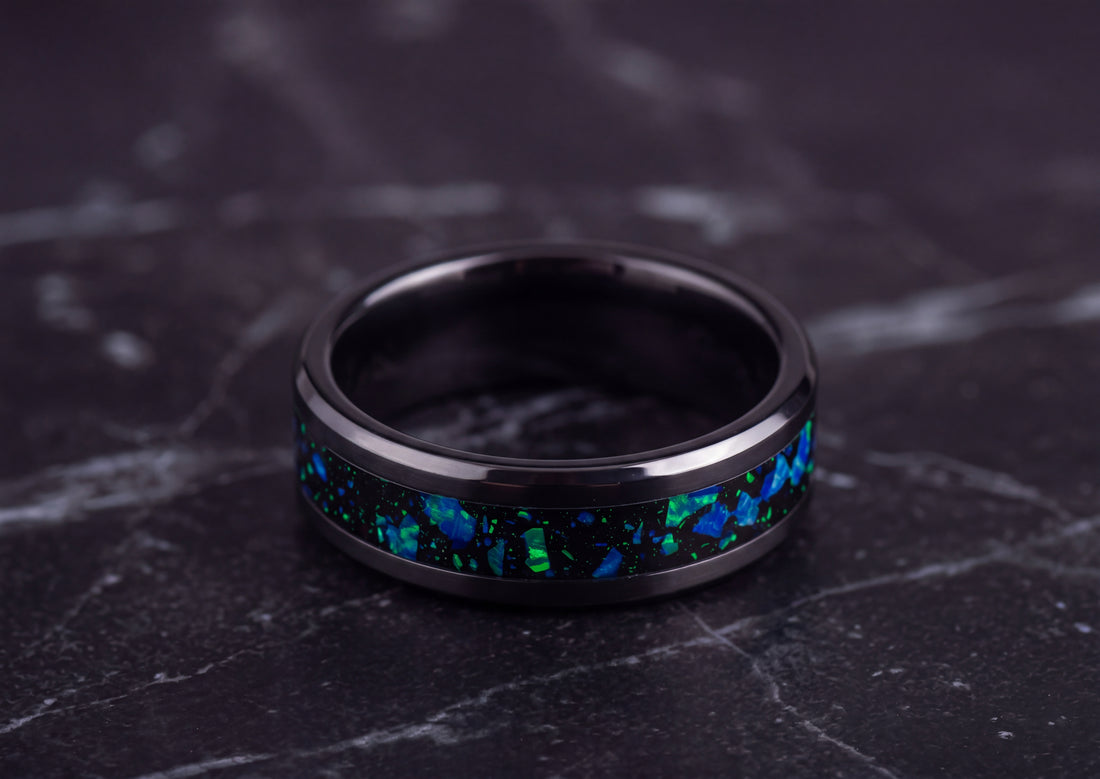 A black metallic ring with a vibrant blue and green iridescent inlay, resting on a dark gray marble surface.