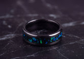 A black metallic ring with a vibrant blue and green iridescent inlay, resting on a dark gray marble surface.