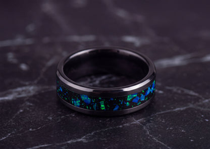 A black metallic ring with a vibrant blue and green iridescent inlay, resting on a dark gray marble surface.