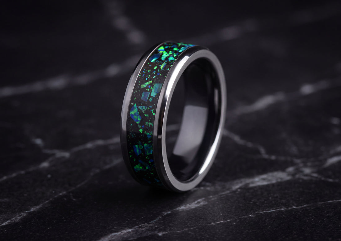 A polished dark ceramic ring with a vibrant green and blue iridescent inlay, displayed on a dark marble surface.