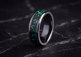 A polished dark ceramic ring with a vibrant green and blue iridescent inlay, displayed on a dark marble surface.