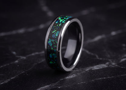 A polished dark ceramic ring with a vibrant green and blue iridescent inlay, displayed on a dark marble surface.