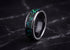 A polished dark ceramic ring with a vibrant green and blue iridescent inlay, displayed on a dark marble surface.