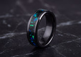 A black metal ring with a vibrant blue and green opal inlay, displayed on a dark marble surface.