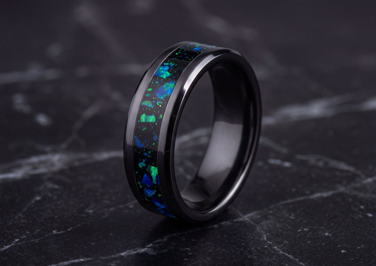 A black metal ring with a vibrant blue and green opal inlay, displayed on a dark marble surface.
