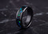 A black metal ring with a vibrant blue and green opal inlay, displayed on a dark marble surface.