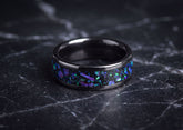 A black ceramicring band featuring a vibrant iridescent inlay of purple, blue, and green opal-like flakes, photographed on a dark marble surface.