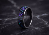 A black ceramic ring with a vibrant purple and blue opal inlay, displayed on a dark marble surface.