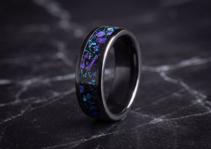 A black ceramic ring with a vibrant purple and blue opal inlay, displayed on a dark marble surface.
