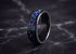 A black ceramic ring with a vibrant purple and blue opal inlay, displayed on a dark marble surface.