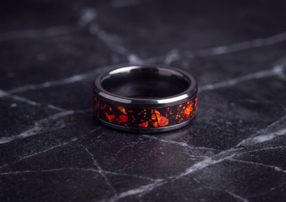 A black ceramic ring band featuring a vibrant orange and red fiery inlay, resting on a dark gray marble surface.