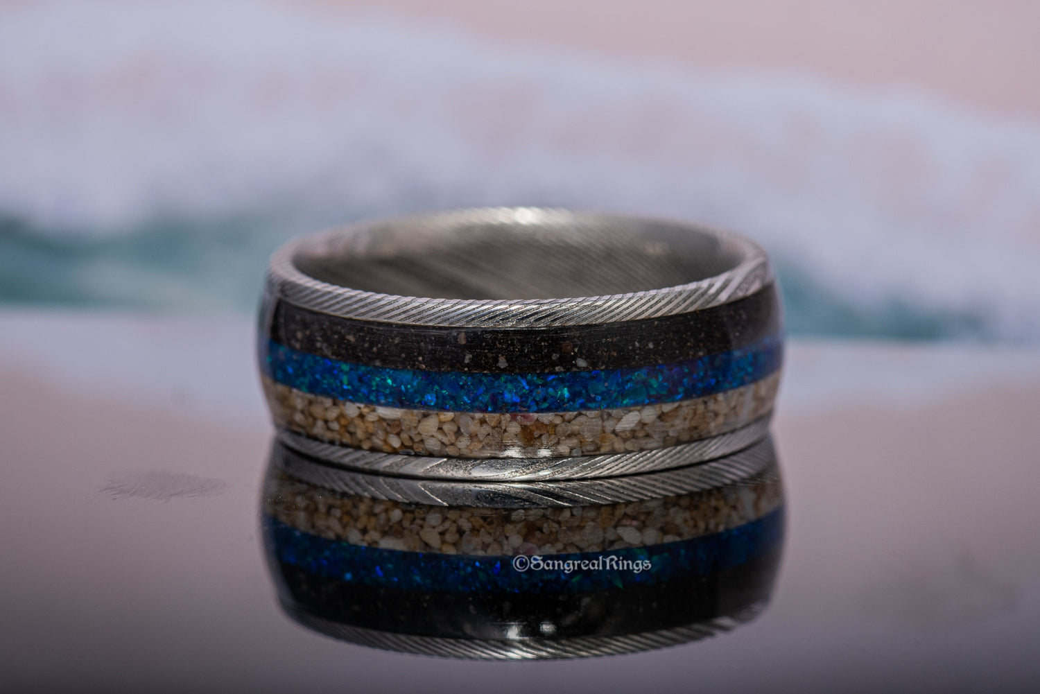 Damascus steel sand ring by Sangreal Rings with sand and blue inlay on reflective surface