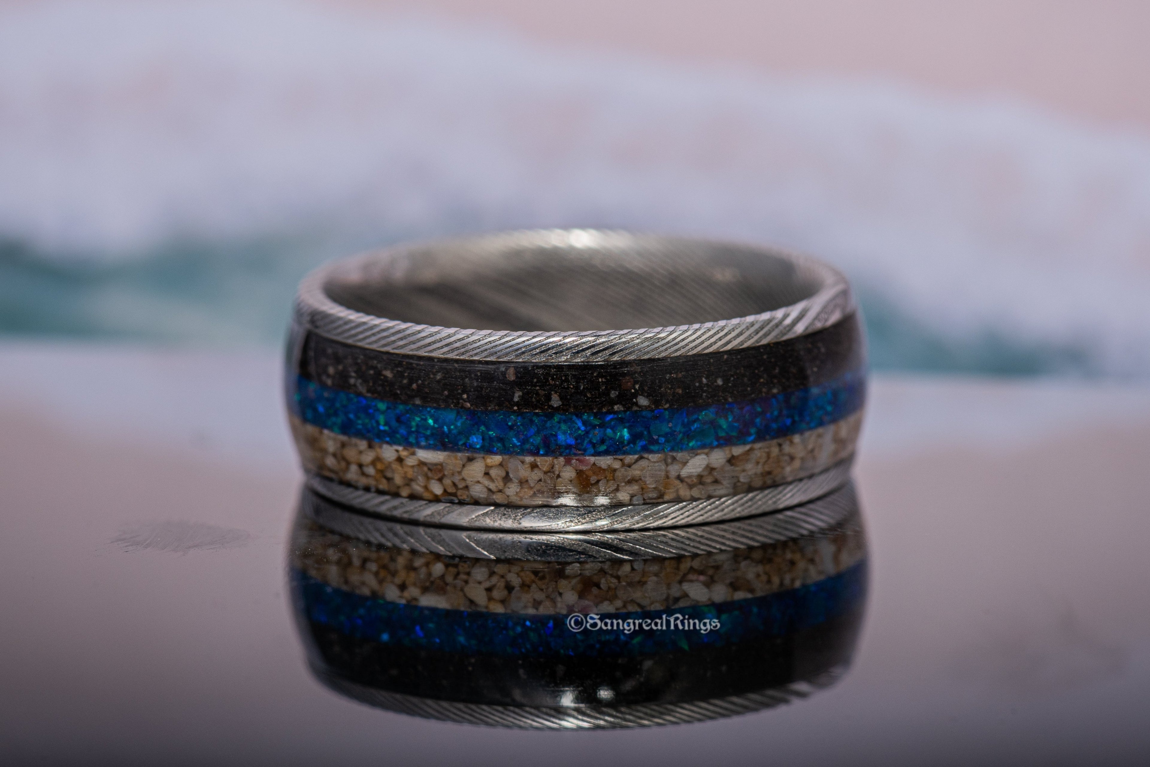 Damascus steel sand ring by Sangreal Rings with sand and blue inlay on reflective surface