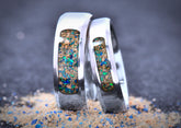 Two silver colored Titanium wedding rings with embedded colorful opal and stones on a sandy surface.