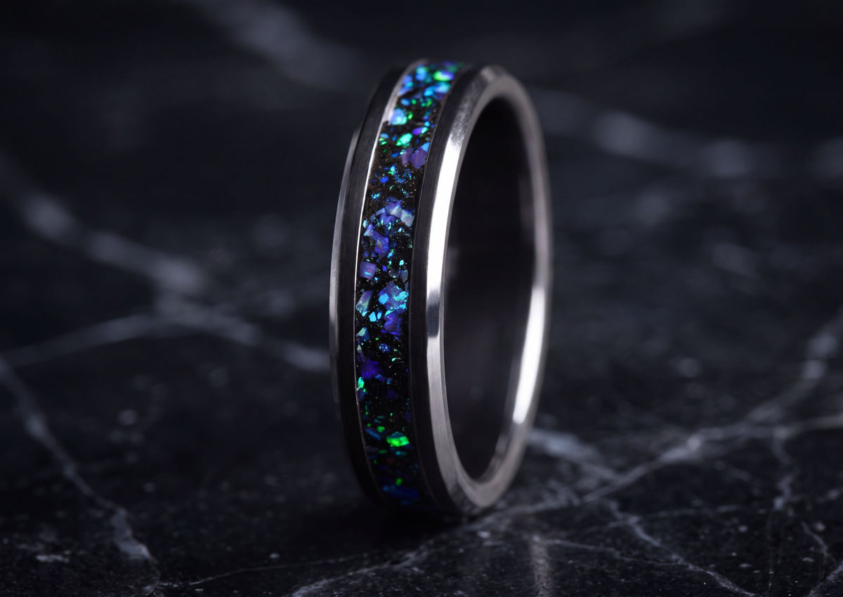 A polished dark metallic ring with a vibrant inlay of iridescent blue, green, and purple opal flakes, set against a dark marble background.