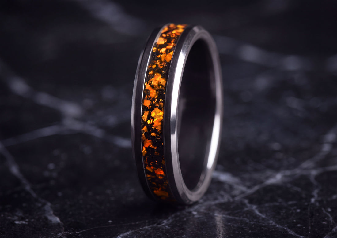 A polished dark metallic ring with a vibrant orange and black textured inlay, standing upright on a dark marble surface.