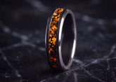 A polished dark metallic ring with a vibrant orange and black textured inlay, standing upright on a dark marble surface.