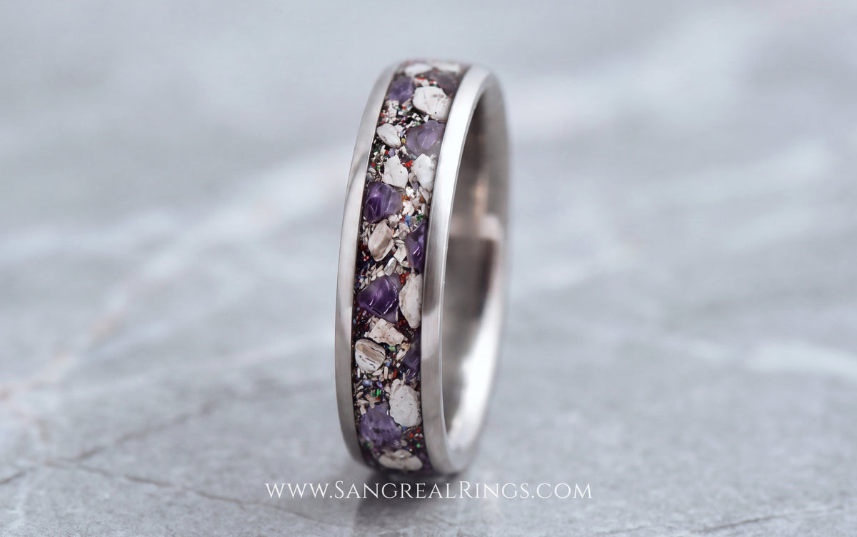 A silver titanium band ring featuring a textured inlay of purple amethyst and cremation ashes, shown on a gray stone surface.