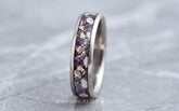 A silver titanium band ring featuring a textured inlay of purple amethyst and cremation ashes, shown on a gray stone surface.