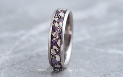 A silver titanium band ring featuring a textured inlay of purple amethyst and cremation ashes, shown on a gray stone surface.
