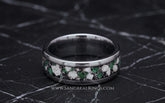 A silver-toned titanium band ring with a channel inlay of green emarald and white cremation ashes, displayed on a dark marble surface. 