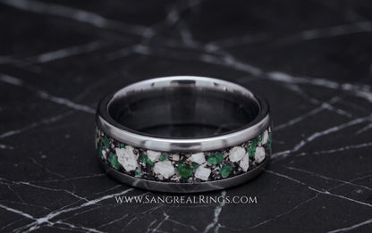 A silver-toned titanium band ring with a channel inlay of green emarald and white cremation ashes, displayed on a dark marble surface. 