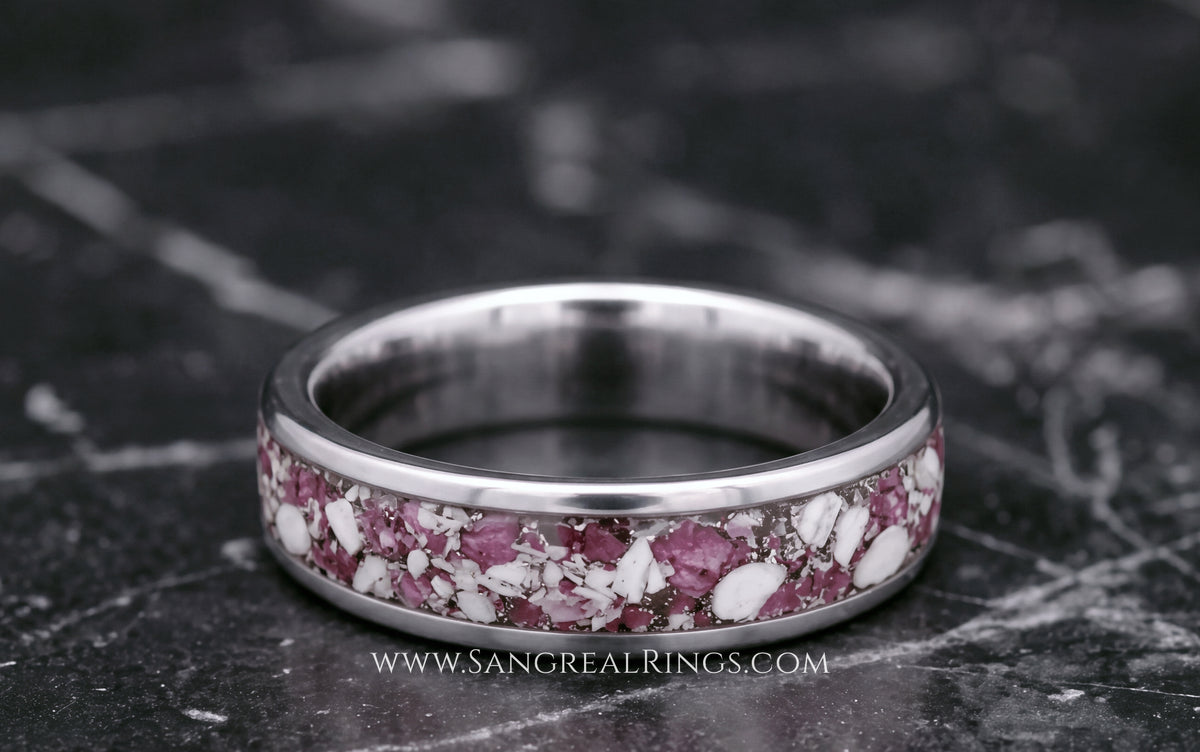 A silver titanium band ring featuring a textured inlay of pink ruby and white cremation ashes resting on a dark gray marble surface.