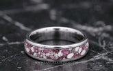 A silver titanium band ring featuring a textured inlay of pink ruby and white cremation ashes resting on a dark gray marble surface.