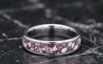 A silver titanium band ring featuring a textured inlay of pink ruby and white cremation ashes resting on a dark gray marble surface.