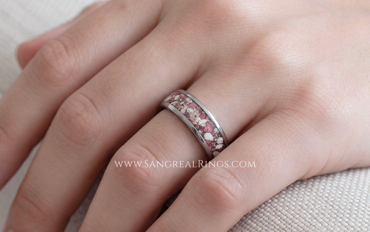 A close-up of a hand wearing a silver band ring with a textured, speckled pink ruby and white cremation ashes inlay. 