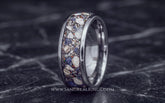 A polished silver-toned titanium ring with a textured inlay of small cremation ashes shards and blue sapphire shards, standing upright on a dark marble surface. 