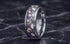 A polished silver-toned titanium ring with a textured inlay of small cremation ashes shards and blue sapphire shards, standing upright on a dark marble surface. 
