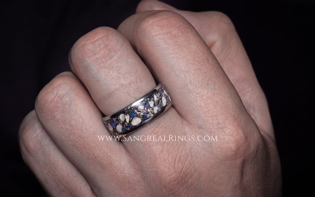A close-up of a hand wearing a titanium silver band ring inlay filled with cremation ashesand dark blue sapphire stones, set against a dark background